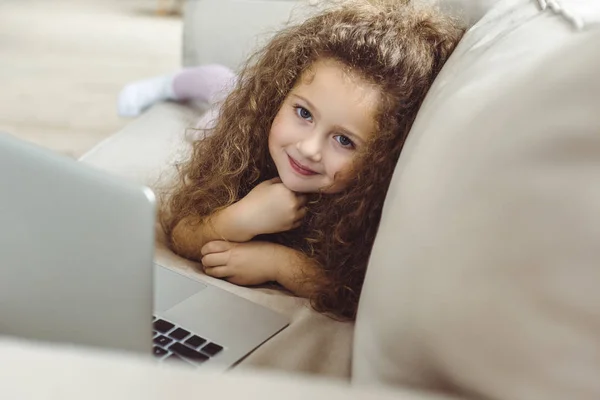 Niño rizado sonriente usando el ordenador portátil y mirando a la cámara - foto de stock