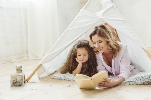 Feliz mãe e filha lendo livro juntos na criança wigwam — Fotografia de Stock