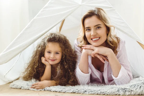 Sonriente mamá y feliz rizado hija acostado en niño wigwam - foto de stock