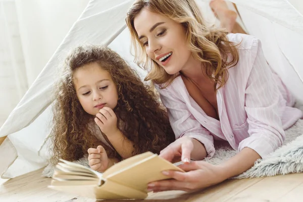 Beautiful mother and daughter reading book in kid wigwam — Stock Photo