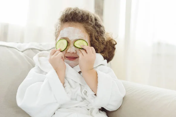 Enfant en peignoir de bain avec des tranches de concombre sur les yeux et masque facial sur le visage — Photo de stock