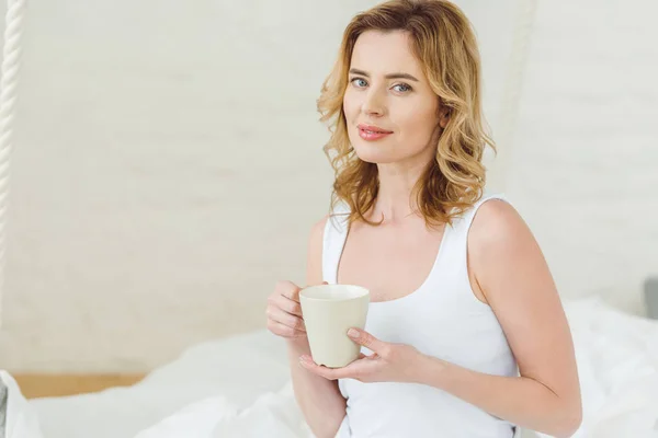 Mujer rubia sonriente con taza de café en el dormitorio por la mañana - foto de stock