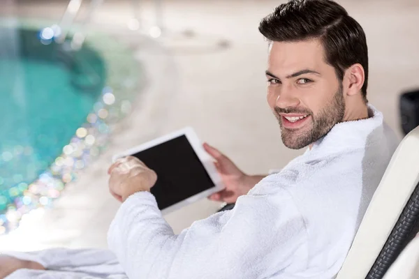 Schöner junger Mann im Bademantel mit digitalem Tablet und lächelnd in die Kamera im Wellness-Center — Stockfoto