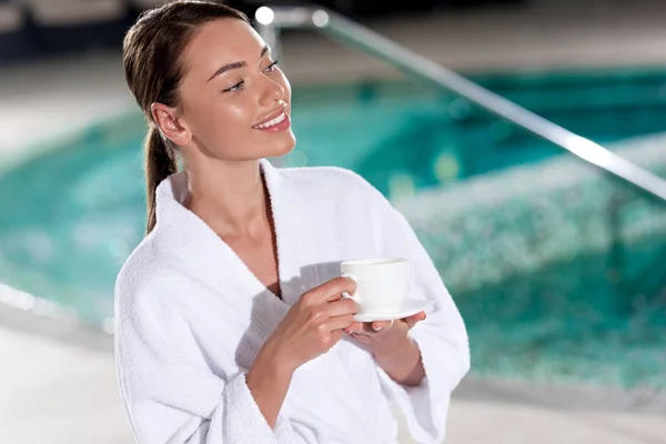 Hermosa mujer joven en albornoz sosteniendo taza de café cerca de la piscina - foto de stock