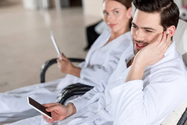 Jovem casal em roupões de banho usando dispositivos digitais enquanto descansam juntos no centro de spa — Fotografia de Stock