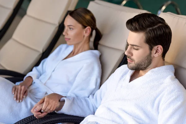 Junges Paar in Bademänteln hält Händchen, während es sich im Wellness-Center ausruht — Stockfoto