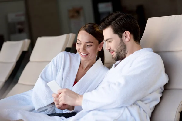 Glückliches junges Paar in Bademänteln mit Smartphone zusammen beim Ausruhen im Wellness-Center — Stockfoto