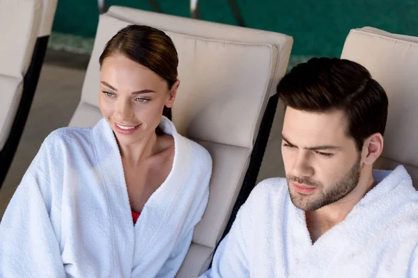 Feliz pareja joven en albornoces sonriendo y mirando hacia otro lado en el centro de spa - foto de stock