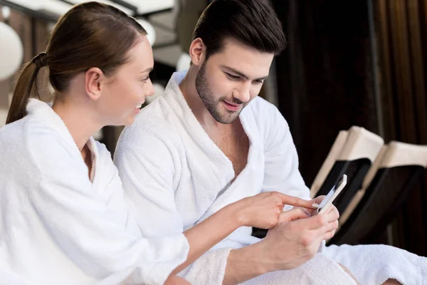 Smiling young couple in bathrobes using smartphone together in spa center — Stock Photo