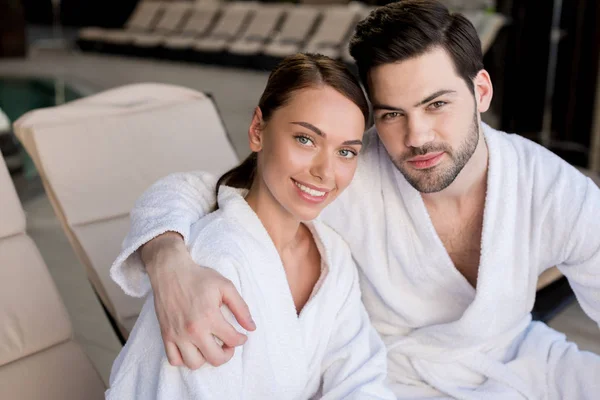 Hermosa pareja joven en albornoces abrazando y sonriendo a la cámara en el centro de spa - foto de stock