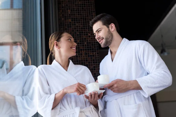 Pareja en albornoces sosteniendo tazas de té y sonriéndose en el centro de spa - foto de stock