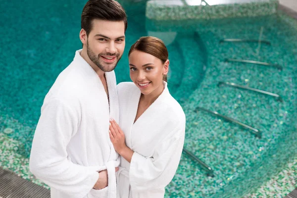 Hermosa joven pareja sonriente en albornoces de pie cerca de la piscina en el centro de spa - foto de stock