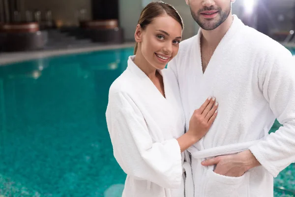 Tiro recortado de casal jovem sorridente em roupões de banho de pé perto da piscina no centro de spa — Fotografia de Stock