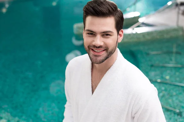 Joven guapo en albornoz sonriendo a la cámara mientras está de pie en el centro de spa - foto de stock