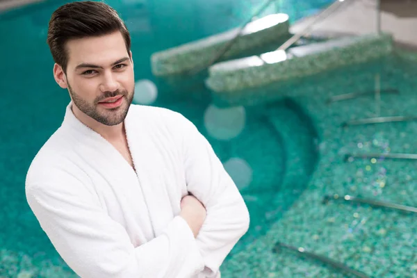 Jovem bonito em roupão de banho de pé com braços cruzados e olhando para a câmera no centro de spa — Fotografia de Stock