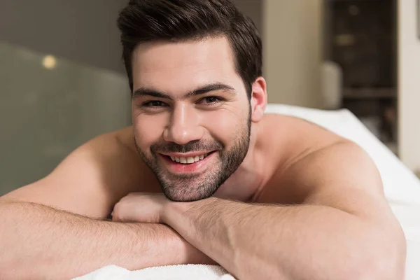 Beau jeune homme barbu souriant à la caméra tout en étant couché dans le salon de spa — Photo de stock