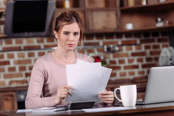 Retrato de la mujer mirando a la cámara mientras hace papeleo en la mesa en casa - foto de stock