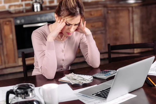 Retrato de la mujer frustrada sentada en la mesa con dinero, ordenador portátil y papeles en casa, concepto de problemas financieros - foto de stock