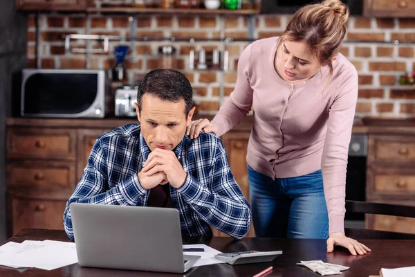 Portrait of woman cheering up upset husband that sitting at table with laptop at home, financial problems concept — Stock Photo