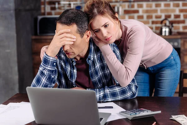 Retrato de la mujer abrazando marido molesto que se sienta a la mesa con el ordenador portátil en casa, concepto de problemas financieros - foto de stock