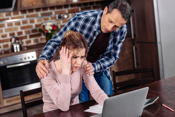 Homem animando esposa chateada que sentado à mesa com laptop em casa, problemas financeiros conceito — Fotografia de Stock