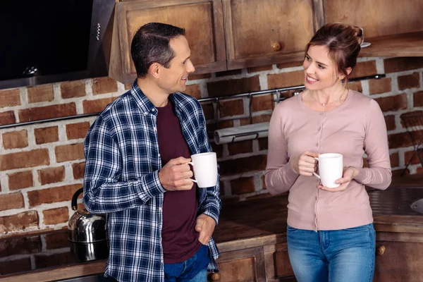 Портрет улыбающейся жены и мужа с чашками кофе разговаривающих на кухне дома — стоковое фото