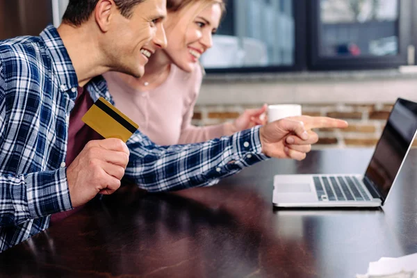 Счастливая пара с кредитной картой и чашкой кофе, глядя на экран ноутбука дома — стоковое фото