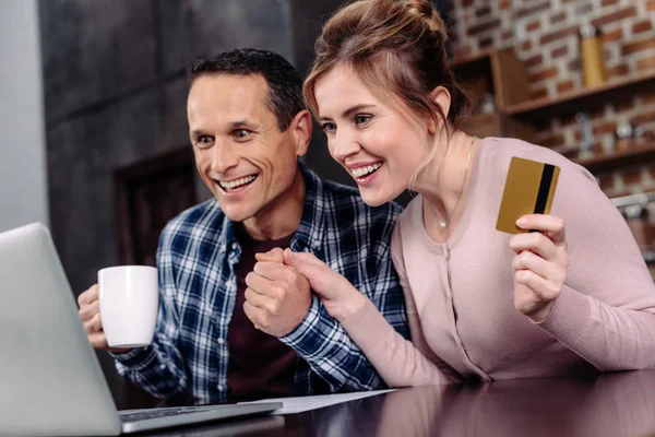 Счастливая пара с кредитной картой и чашкой кофе, глядя на экран ноутбука дома — стоковое фото