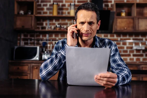 Retrato del hombre sorprendido hablando en el teléfono inteligente mientras mira el papel en la mano en casa - foto de stock