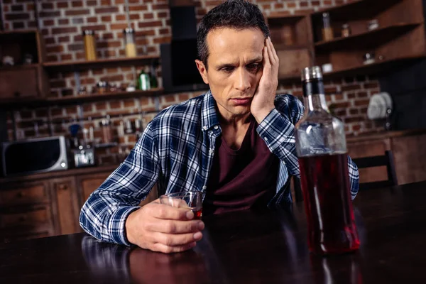 Bouleversé homme assis à table avec bouteille et verre d'alcool à la maison — Photo de stock