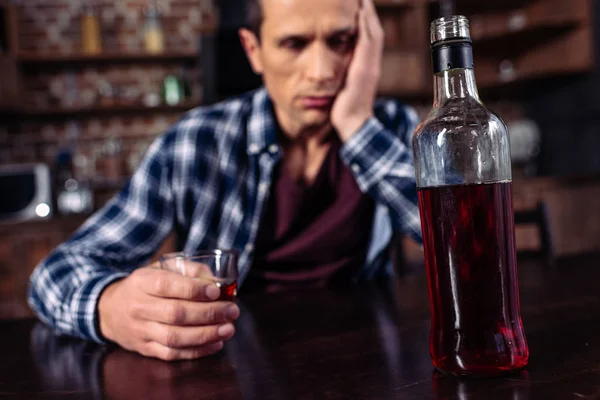 Foco seletivo de homem deprimido sentado à mesa com garrafa e copo de álcool em casa — Fotografia de Stock
