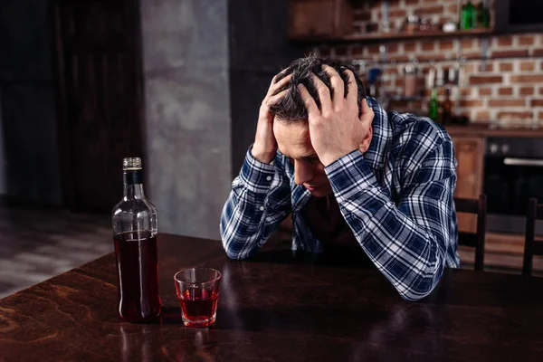 Uomo ubriaco seduto a tavola con bottiglia e bicchiere di alcol a casa — Foto stock