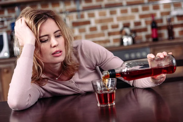 Mujer vertiendo alcohol en el vidrio mientras está sentado en la mesa en la cocina - foto de stock