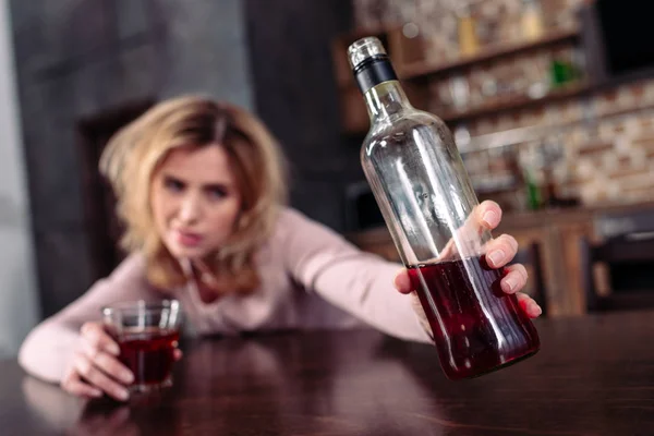 Selektiver Fokus der betrunkenen Frau, die eine Flasche Alkohol nimmt, während sie zu Hause am Tisch sitzt — Stockfoto