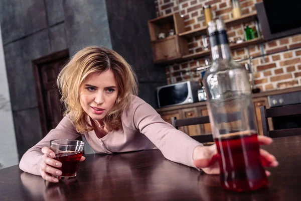 Selective focus of drunk woman taking bottle of alcohol while sitting at table at home — Stock Photo