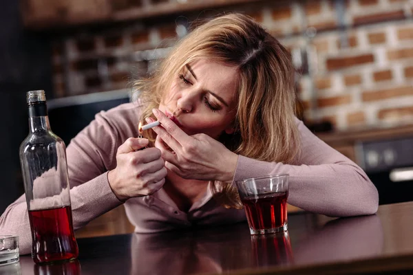 Retrato de la mujer fumando cigarrillo mientras está sentada en la mesa con un vaso de alcohol en casa - foto de stock