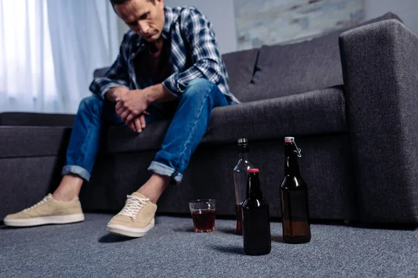 Мужчина смотрит на алкоголь на полу, сидя дома на диване — стоковое фото