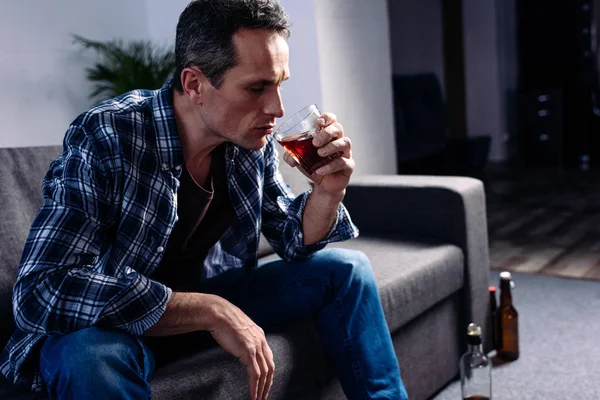 Vista lateral del hombre con vaso de alcohol sentado en el sofá en casa - foto de stock