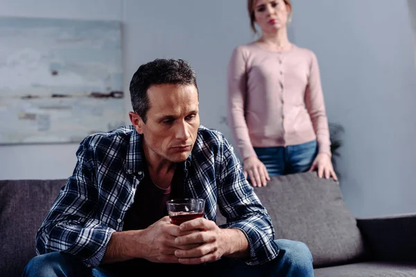 Мужчина со стаканом алкоголя сидит на диване, в то время как жена стоит позади дома — стоковое фото