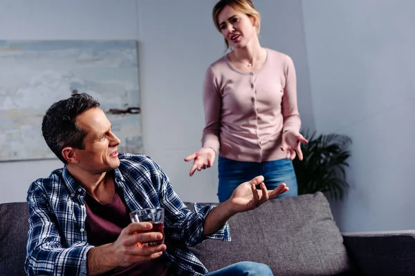 Homem com copo de álcool sentado no sofá, enquanto discutindo com a esposa atrás em casa — Fotografia de Stock