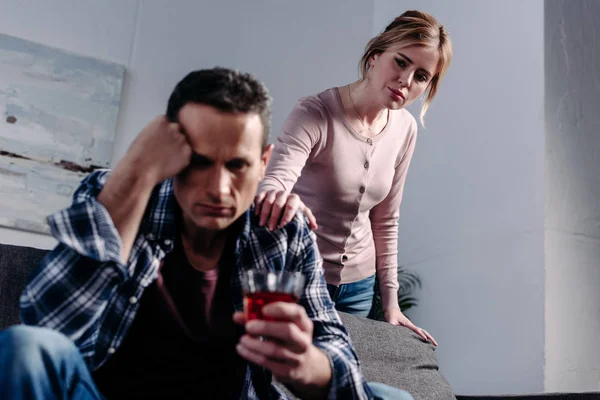 Man with glass of alcohol sitting on sofa while wife standing behind at home — Stock Photo