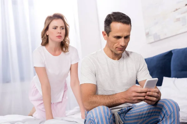 Confused wife looking at husband using smartphone while sitting on bed at home — Stock Photo