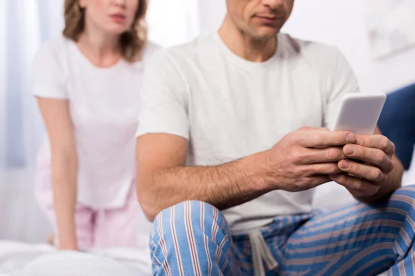 Vista parcial del hombre usando el teléfono inteligente con la esposa de pie detrás en casa - foto de stock