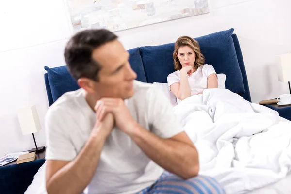 Selective focus of upset man sitting on bed with wife behind at home, relationship difficulties concept — Stock Photo
