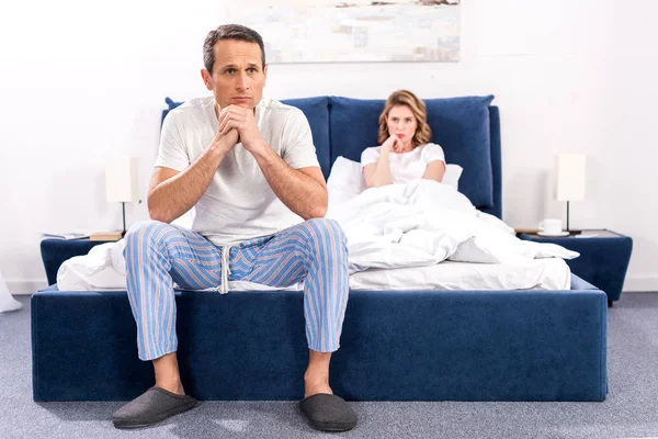 Chateado homem sentado na cama com a esposa atrás em casa, relacionamento dificuldades conceito — Fotografia de Stock