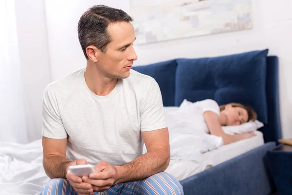 Enfoque selectivo del hombre usando el teléfono inteligente mientras su esposa duerme en la cama en casa - foto de stock