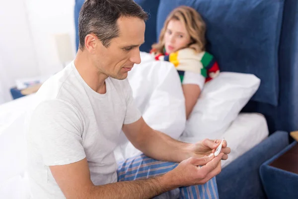 Hombre mirando el termómetro en las manos con la esposa enferma en la cama cerca - foto de stock