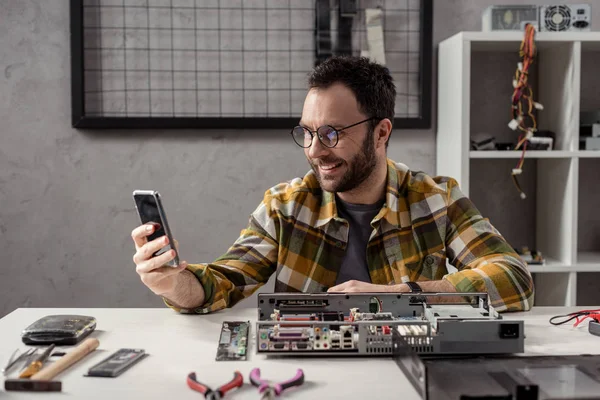 Улыбающийся мужчина использует смартфон, сидя на сломанном компьютере — стоковое фото