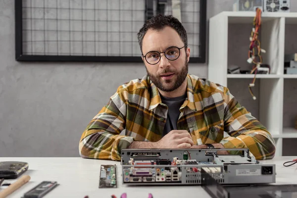 Mann blickt auf Kamera gegen kaputten Computer auf Tisch — Stockfoto