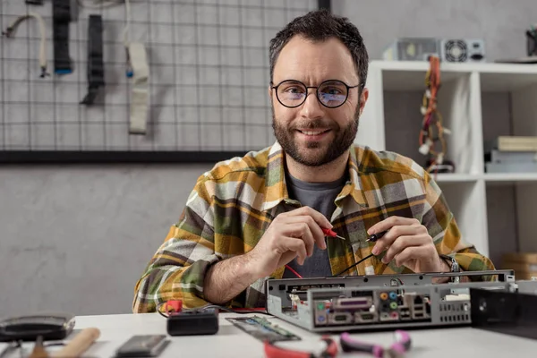 Lächelnder Mann schaut in Kamera, während er Multimeter über kaputtem PC benutzt — Stockfoto